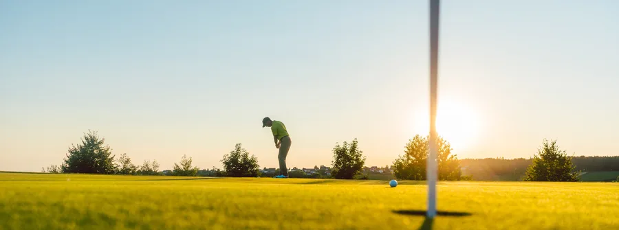 Golfer in a lime-green shirt lines up a putt on a sunlit golf green, with a flagstick casting a long shadow as the sun sets behind trees.