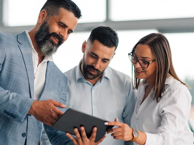 Three colleagues in business attire are looking at a tablet together, smiling, in a modern office with large windows in the background.