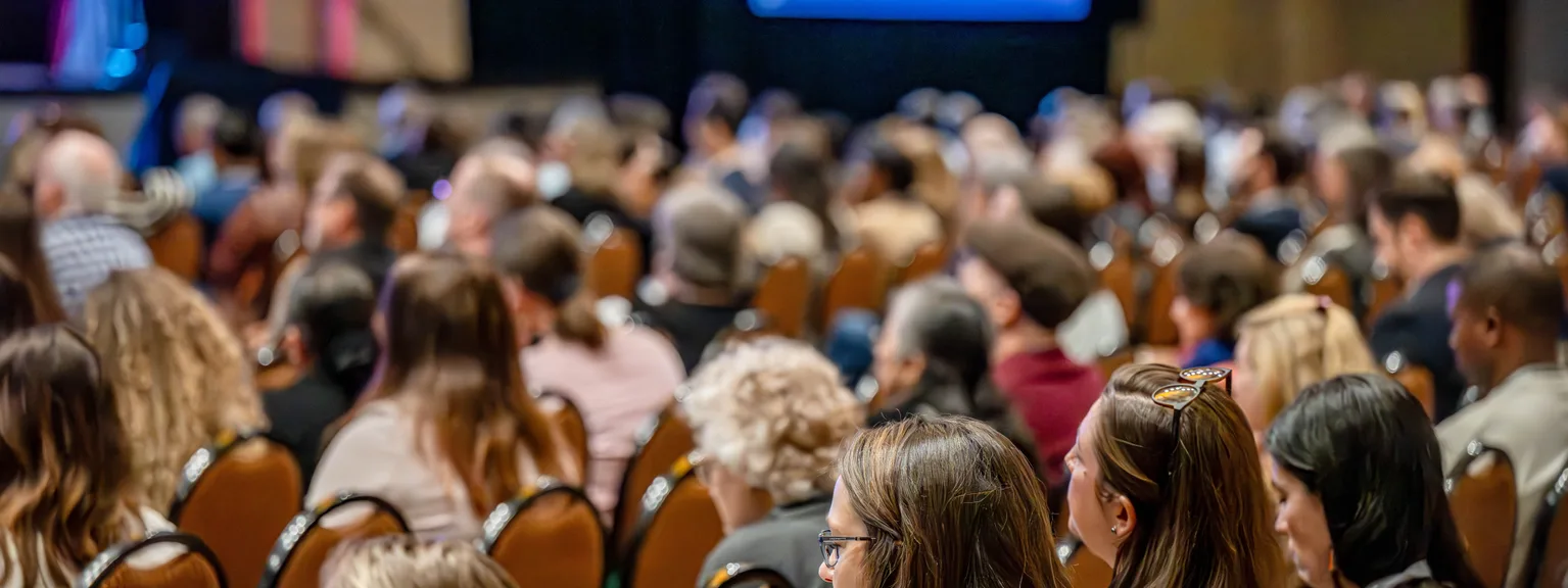 Crowd of people sitting in rows at a conference or seminar, facing a stage with a large screen displaying a blue presentation slide.