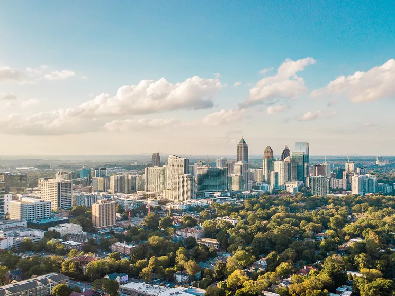 A city skyline with tall buildings and skyscrapers against a blue sky with scattered clouds, overlooking a green residential neighborhood.