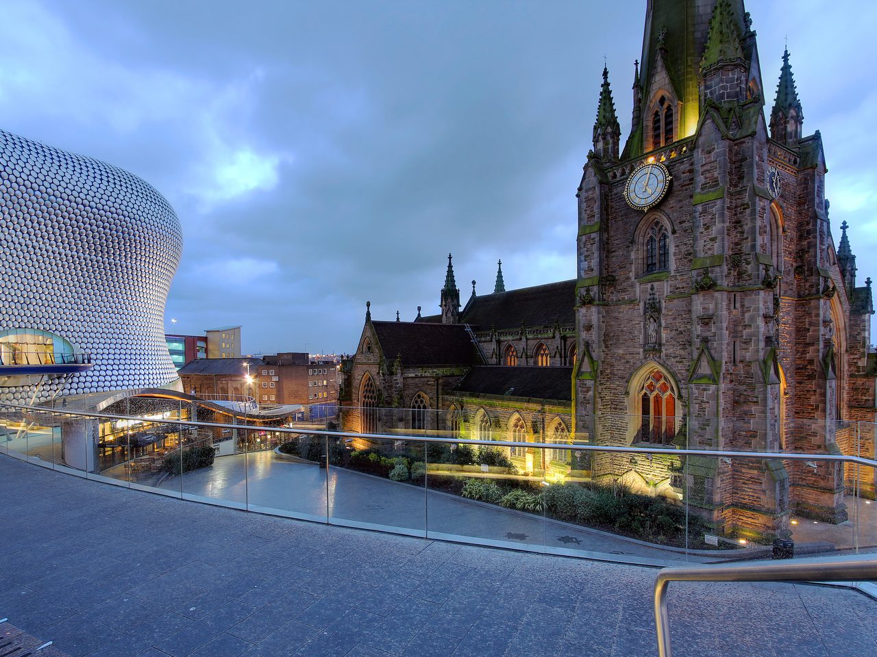 Blue-hour view of a Gothic church with a clock tower beside a curved metallic modern building, with a glass railing and elevated walkway over a plaza.