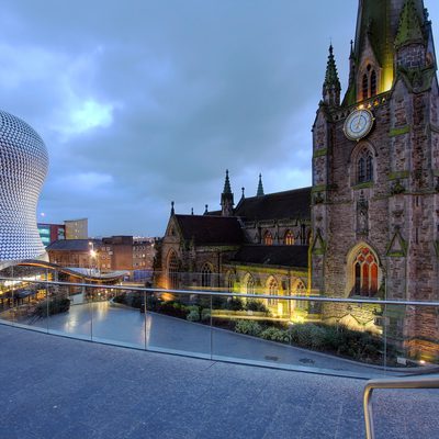 Blue-hour view of a Gothic church with a clock tower beside a curved metallic modern building, with a glass railing and elevated walkway over a plaza.