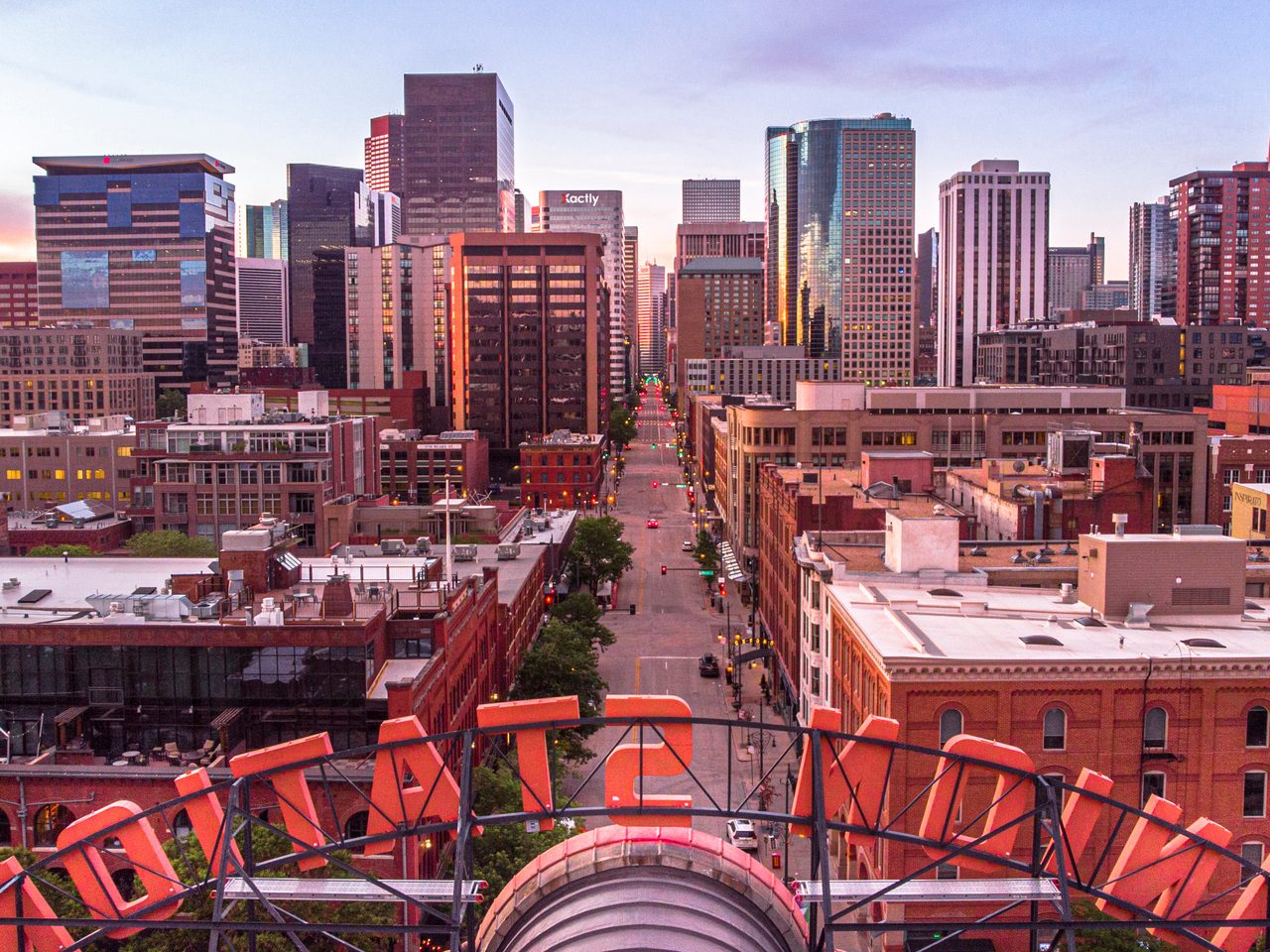 A downtown cityscape at dusk, a main street leads between tall glass towers, with sunset light and red-brick buildings in foreground, from a rooftop.