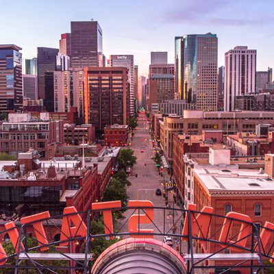 A downtown cityscape at dusk, a main street leads between tall glass towers, with sunset light and red-brick buildings in foreground, from a rooftop.