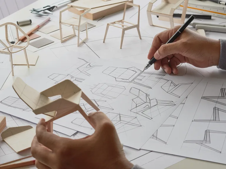 Designer’s hands sketch architectural chair designs on a drafting table, surrounded by wooden chair models, plans and drafting tools.