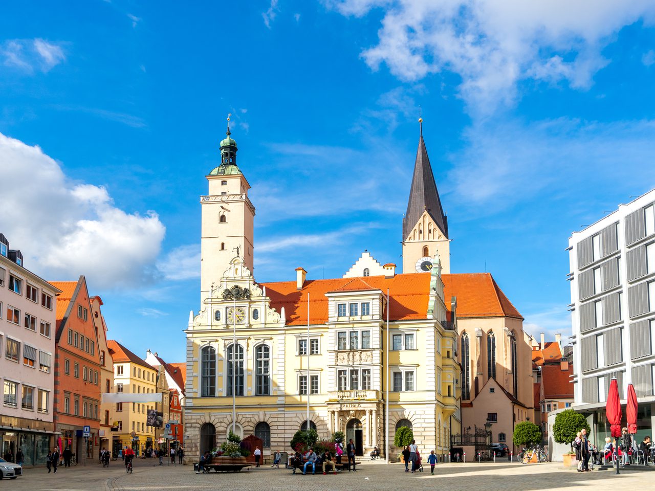 Historic town square with a pale ornate town hall, tall church spire, pastel buildings, and a glass-fronted building on the right; people walking.