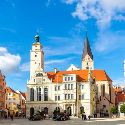 Historic town square with a pale ornate town hall, tall church spire, pastel buildings, and a glass-fronted building on the right; people walking.