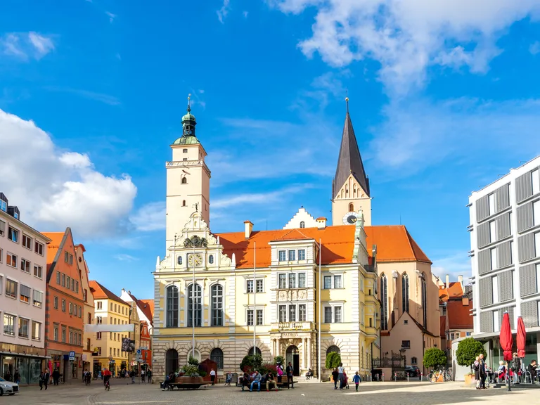 Historic town square with a pale ornate town hall, tall church spire, pastel buildings, and a glass-fronted building on the right; people walking.