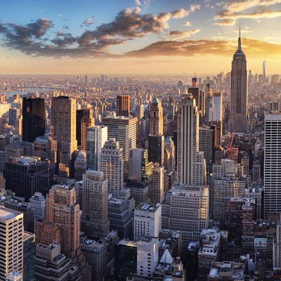 A panoramic view of Manhattan skyline at sunset, featuring tall skyscrapers including Empire State Building, with warm lighting and partly cloudy sky.