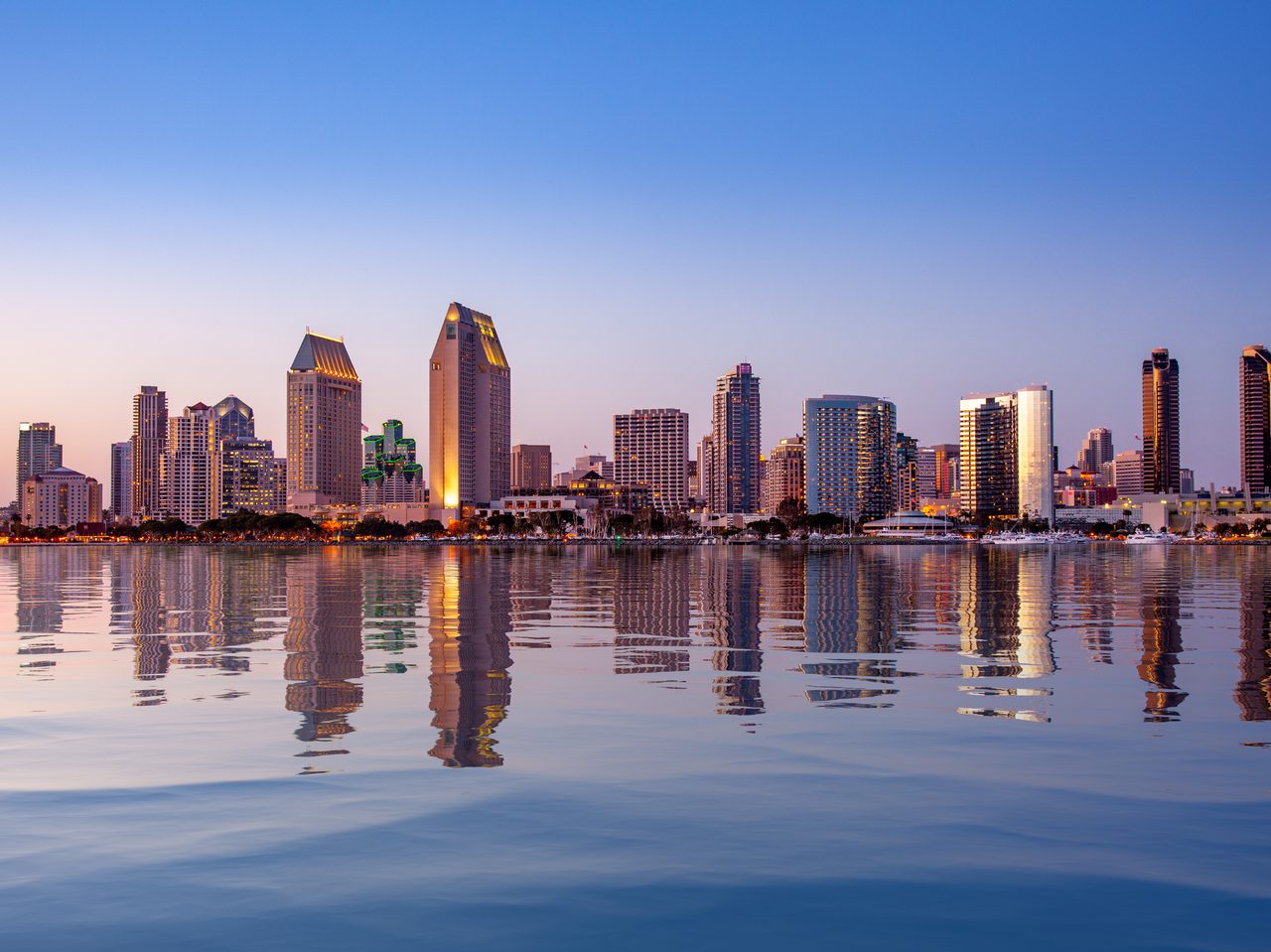 A city skyline reflected in calm water at dusk, featuring tall modern skyscrapers under a clear blue sky.