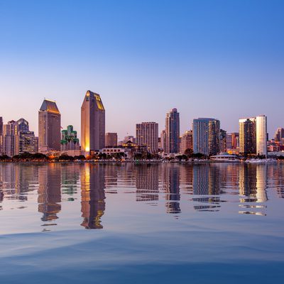 Skyline of modern skyscrapers reflecting in calm water at sunset, with a clear blue sky overhead.