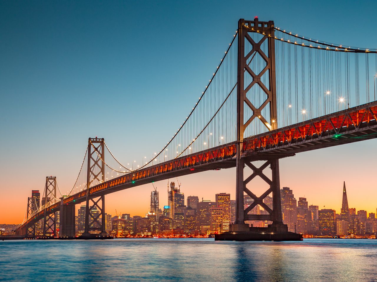 A suspension bridge illuminated at dusk with a city skyline in the background, featuring skyscrapers and a clear gradient sky.