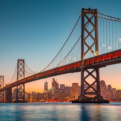 A suspension bridge illuminated at dusk with a city skyline in the background, featuring skyscrapers and a clear gradient sky.