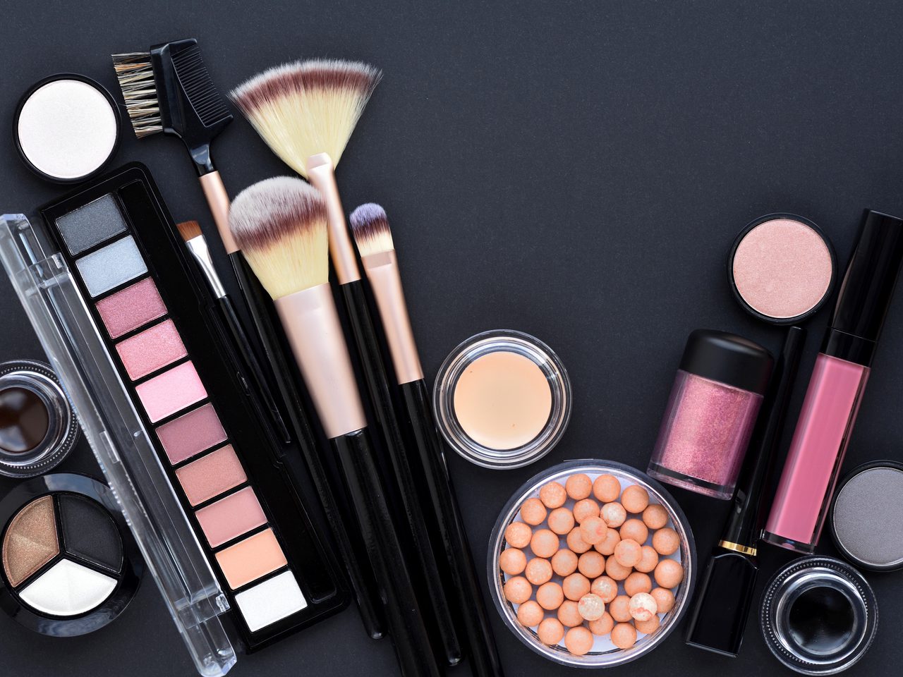 Flat lay of makeup items on a dark surface: eyeshadow palette, brushes, powders, blush beads, and lip gloss, lipstick containers.
