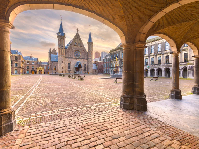 An empty historic square with cobblestone pavement, surrounded by old buildings, viewed through stone archways, at sunrise.
