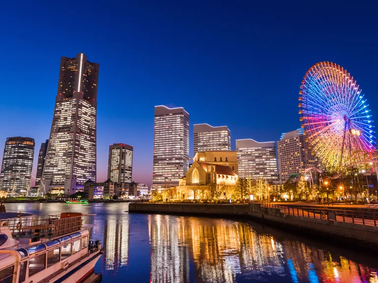 City skyline at dusk with illuminated skyscrapers, a ferris wheel, and reflections on the water in a lively urban setting.
