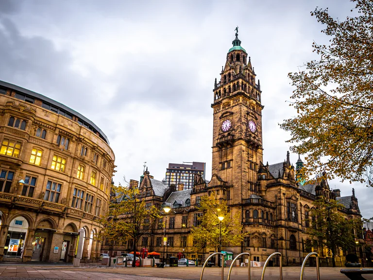 Historischer Rathaus-Turm in Glasgow bei Bewölktem Himmel, umgeben von Bäumen mit Herbstlaub und beleuchteten Laternen.