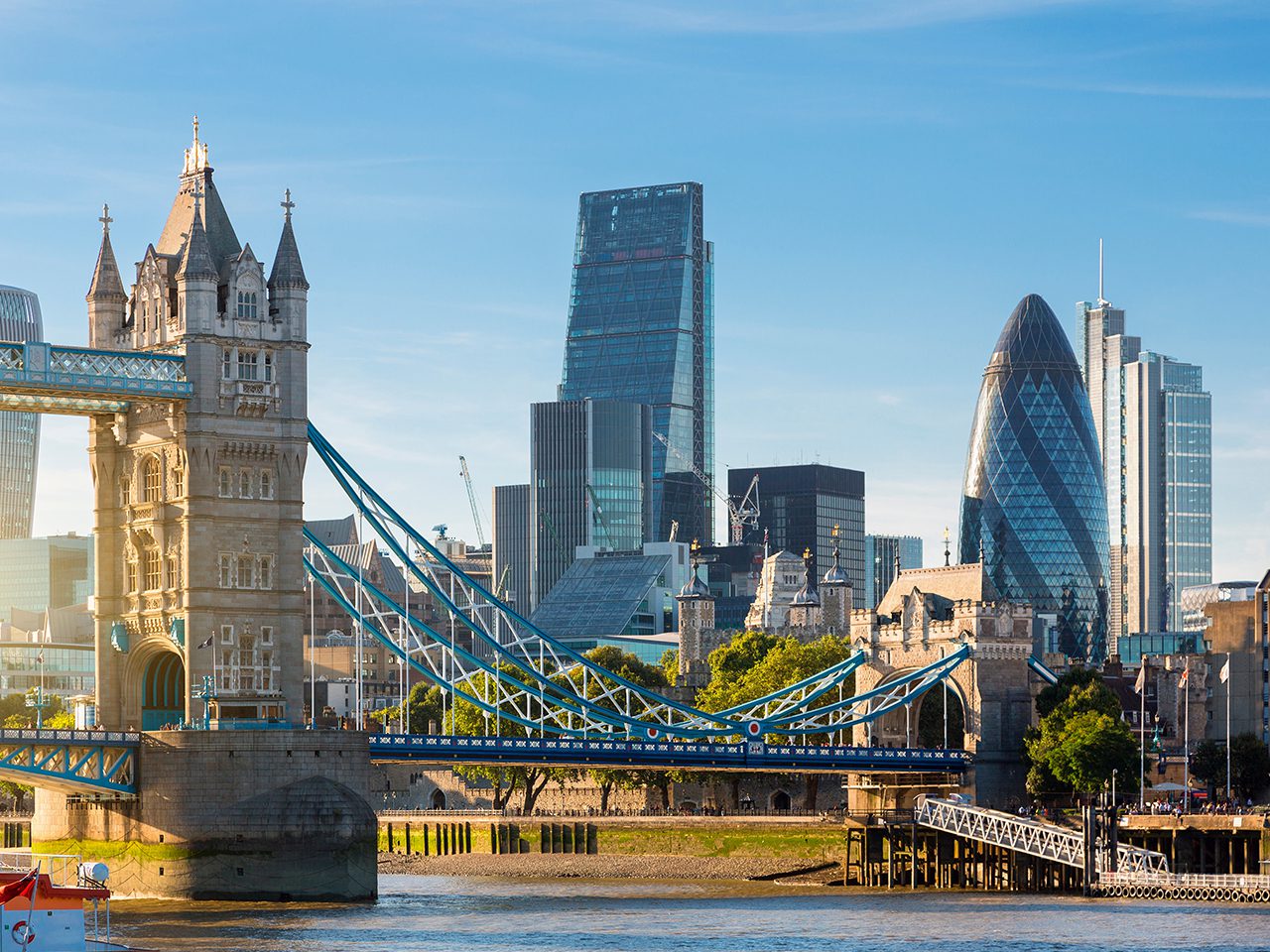 The image shows the London skyline with the Tower Bridge in the foreground and modern skyscrapers in the background under a clear blue sky.