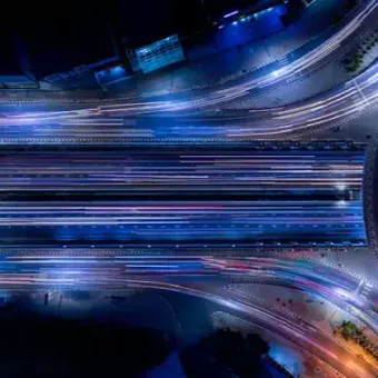 Aerial view of a busy multi-lane highway at night with streaks of traffic lights and illuminated roads, featuring a curved interchange.