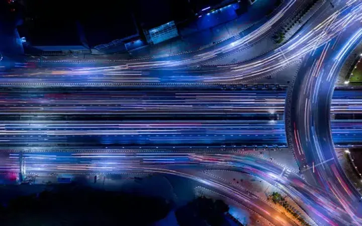 Aerial view of a busy multi-lane highway at night with streaks of traffic lights and illuminated roads, featuring a curved interchange.