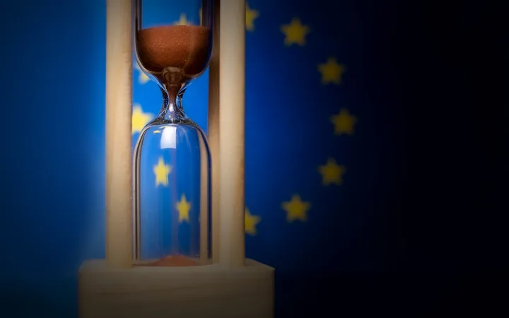 An hourglass with brown sand against a European Union flag background.