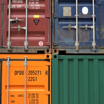 Four colorful shipping containers stacked, with focus on their weathered doors, lock mechanisms, and printed safety and weight information.