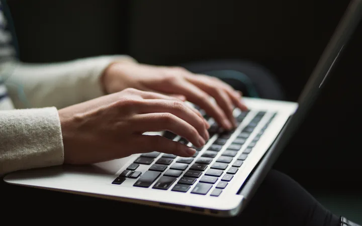 Hands typing on a laptop keyboard, with focus on the hands and keyboard, in a dimly lit environment.