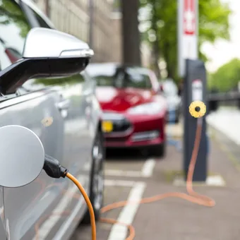 Close-up of an electric car charging at a station on a parking lot, with other cars and trees in the background.
