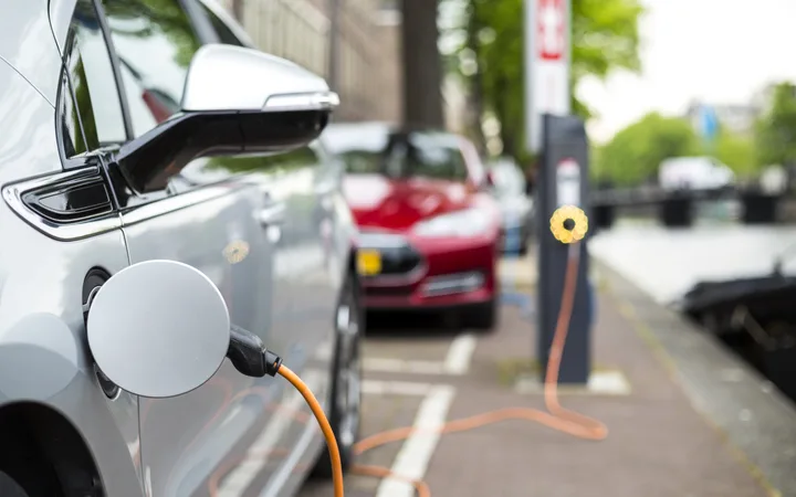 Close-up of an electric car charging at a station on a city street, with a red car and green trees in the background under an overcast sky.