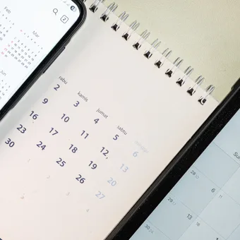Three devices on a beige surface showing calendars: a left smartphone with a yearly calendar, a middle spiral notebook calendar, and a right device with a calendar app.