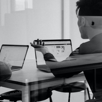 Two people sit at a table working on laptops, one facing left and the other with his back to the camera, in an office environment with natural light.