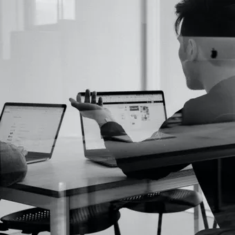 Two people sit at a table working on laptops, one facing left and the other with his back to the camera, in an office environment with natural light.