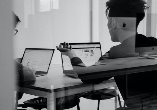 Two people sitting at a table working on laptops, viewed from behind and partly through a glass door, in a modern, well-lit office setting.