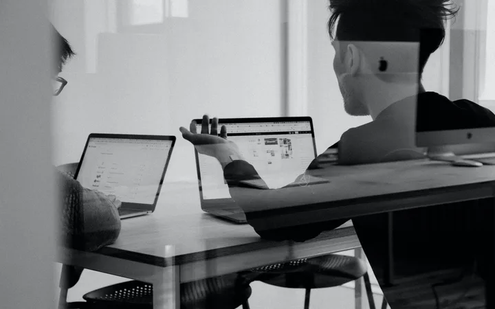 Two people sit at a table working on laptops, one facing left and the other with his back to the camera, in an office environment with natural light.