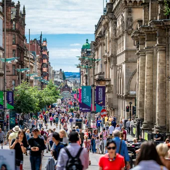 Crowded city street with historic buildings, colorful banners, and diverse pedestrians during daytime in an urban area.
