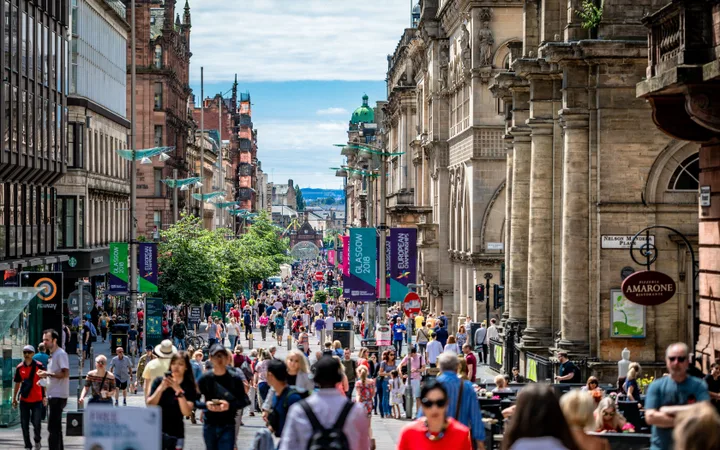 Crowded city street with historic buildings, banners for Glasgow 2018, and pedestrians enjoying a sunny day in an urban shopping district.