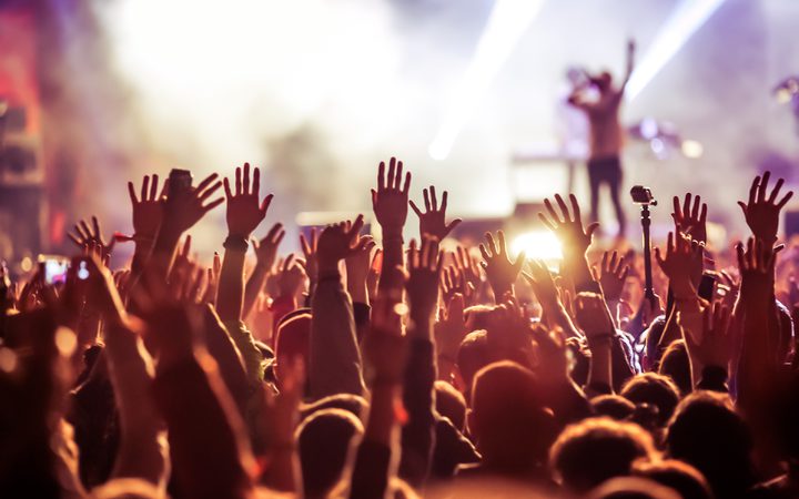 Crowd at concert with hands raised, bright stage lights and a performer in the background, creating an energetic and lively atmosphere.