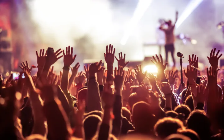 Crowd at an event with raised hands, stage lighting, and a performer in the background creating an energetic atmosphere.