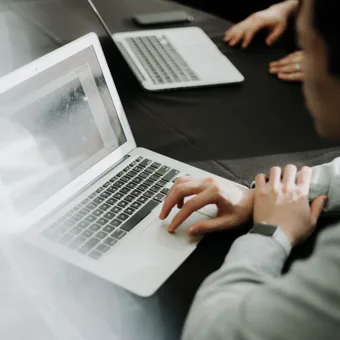 Person using a silver laptop at a conference table with another laptop and a smartphone in the background.