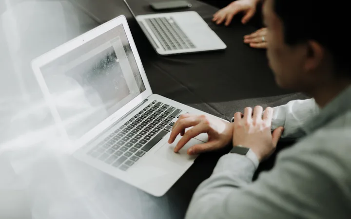 Person using a laptop at a meeting, with another laptop and hand visible on a dark table.