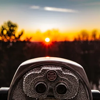 A coin-operated binocular viewer in the foreground with a sunset and silhouetted trees in the background.