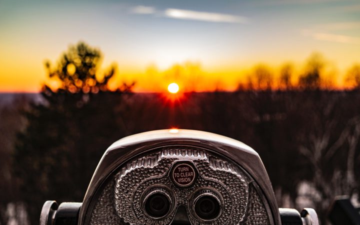 A coin-operated binocular viewer in the foreground with a sunset and silhouetted trees in the background.