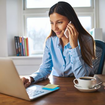 Person in a blue shirt on a video call, sitting at a desk with a laptop, coffee and notes, in a bright home office.