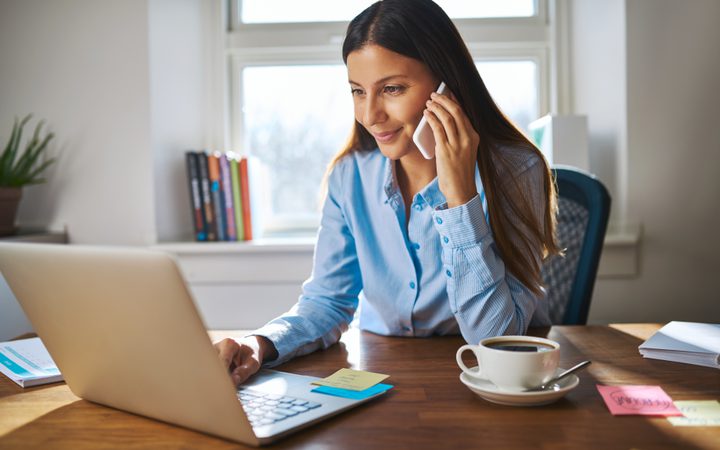 Person in a blue shirt on a video call, sitting at a desk with a laptop, coffee and notes, in a bright home office.