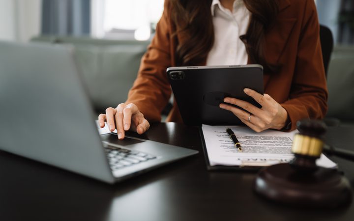 A person in a brown blazer sits at a desk with a laptop, holding a tablet; a printed document with a pen lies on the desk, and a gavel nearby.