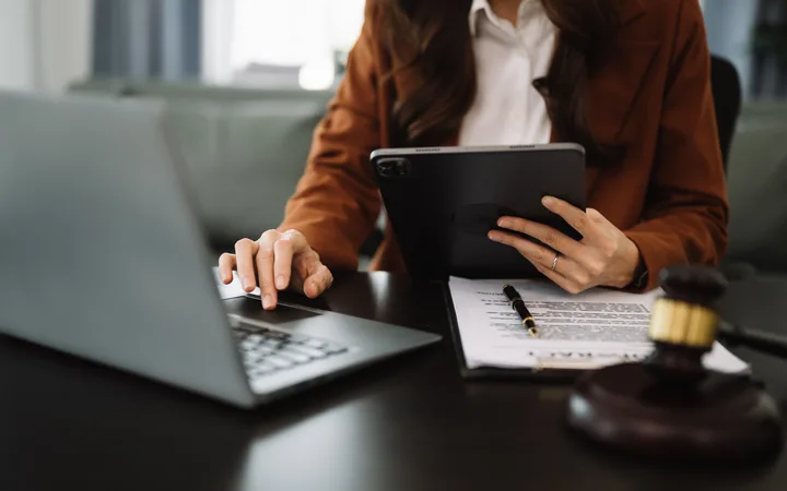 A person in a brown blazer sits at a desk with a laptop, holding a tablet; a printed document with a pen lies on the desk, and a gavel nearby.