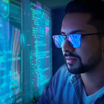 A man wearing glasses looks at multiple computer screens with glowing blue and green data and code, in a dark room, suggesting a high-tech or cybersecurity setting.