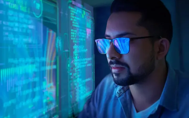 A man wearing glasses looks at multiple computer screens with glowing blue and green data and code, in a dark room, suggesting a high-tech or cybersecurity setting.