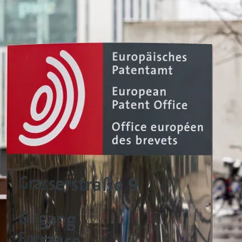 Sign for the European Patent Office in multiple languages with a red and gray color scheme, located near bicycles and a modern building.