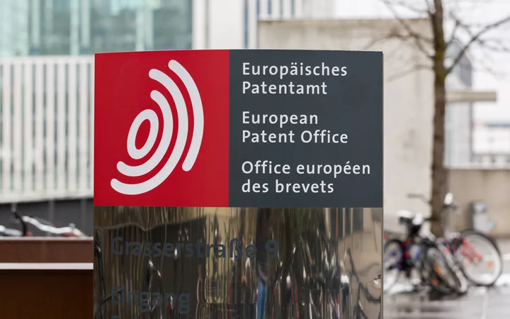Sign for the European Patent Office on a building, with text in German, English, and French, and a red and black background, alongside bicycles parked nearby.